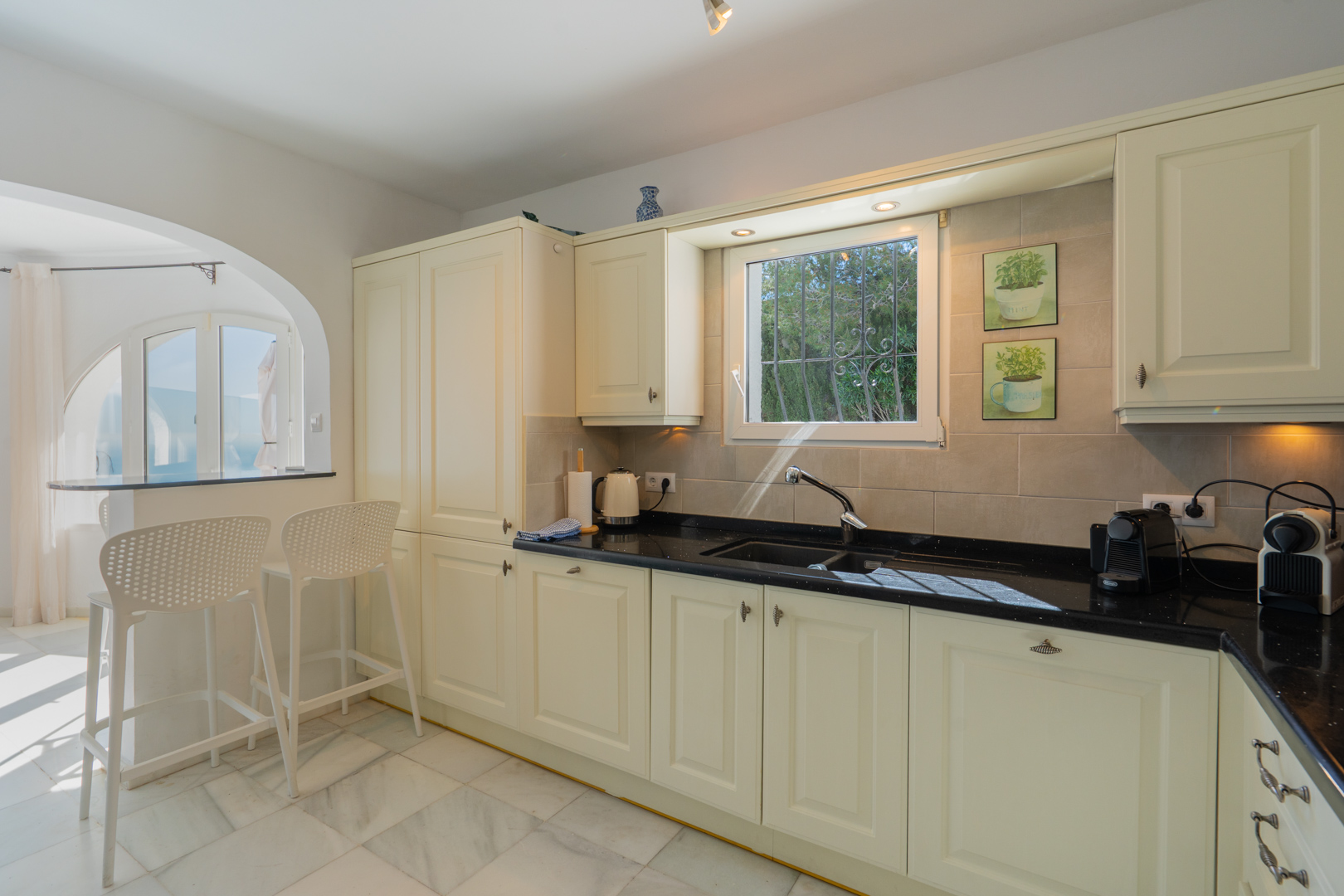 Cocina de estilo tradicional con armarios en tono crema, encimera de granito oscuro y suelos de mármol. Un arco comunica el espacio con la barra de desayuno y el salón.