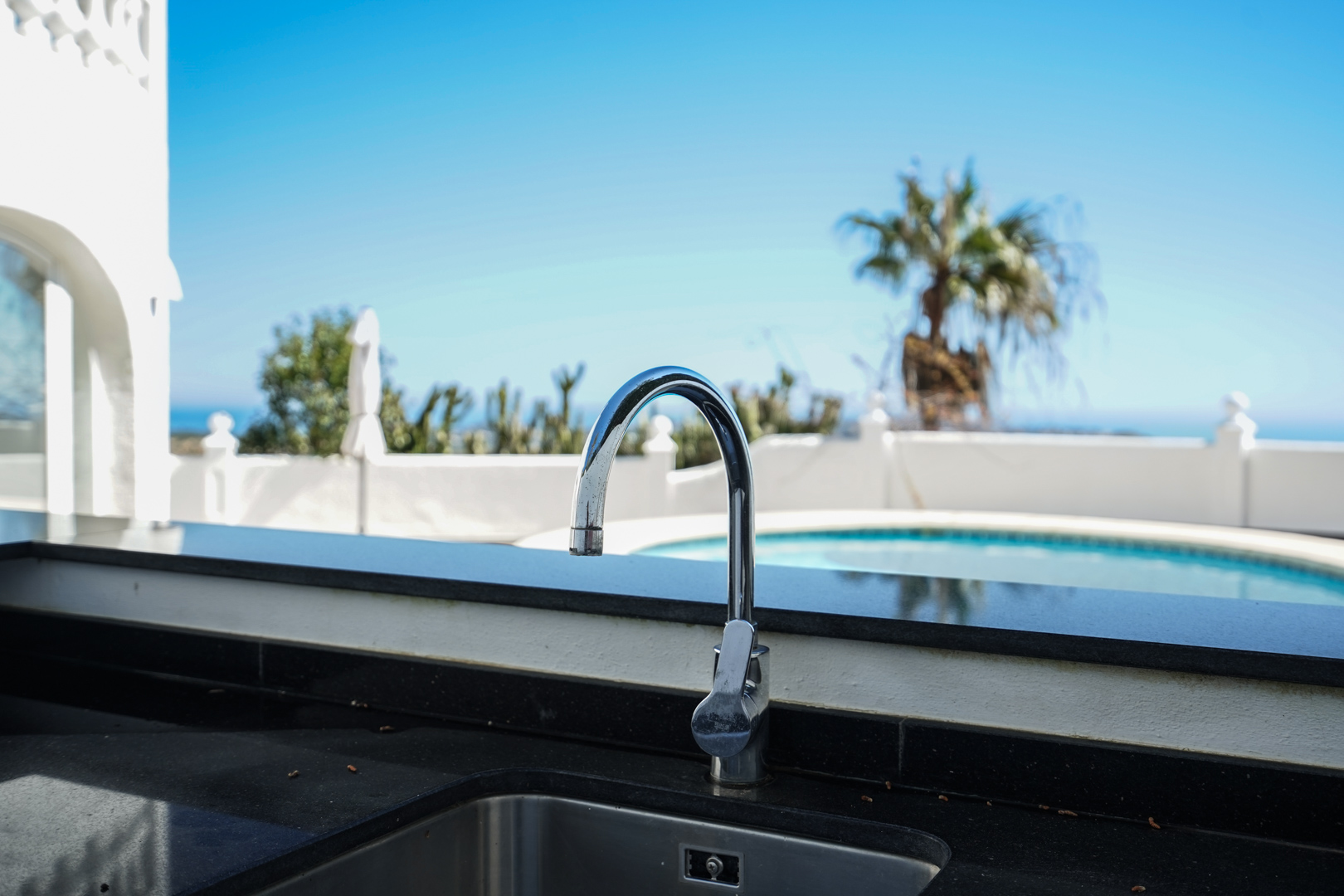 Cocina exterior con encimera de granito negro y grifo cromado, situada frente a la piscina privada con vistas al paisaje mediterráneo.