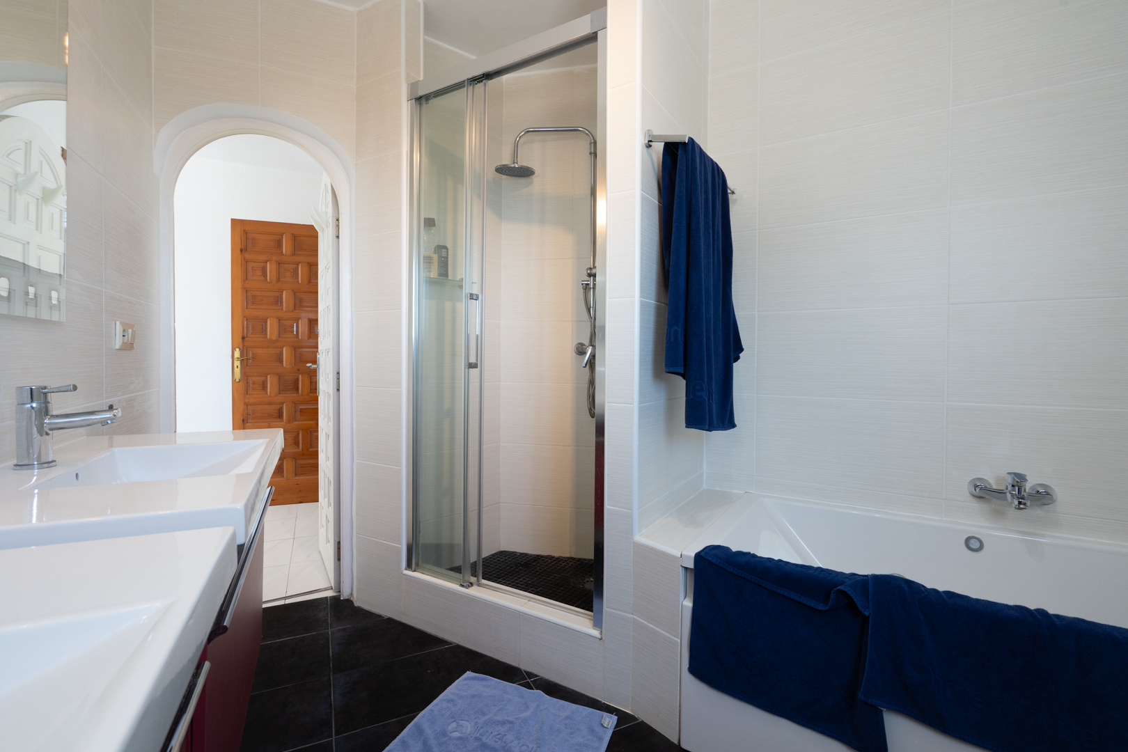 Cuarto de baño equipado con lavabo doble, bañera y ducha independiente con mampara de cristal. Presenta azulejos claros texturizados y un suelo en contraste oscuro.