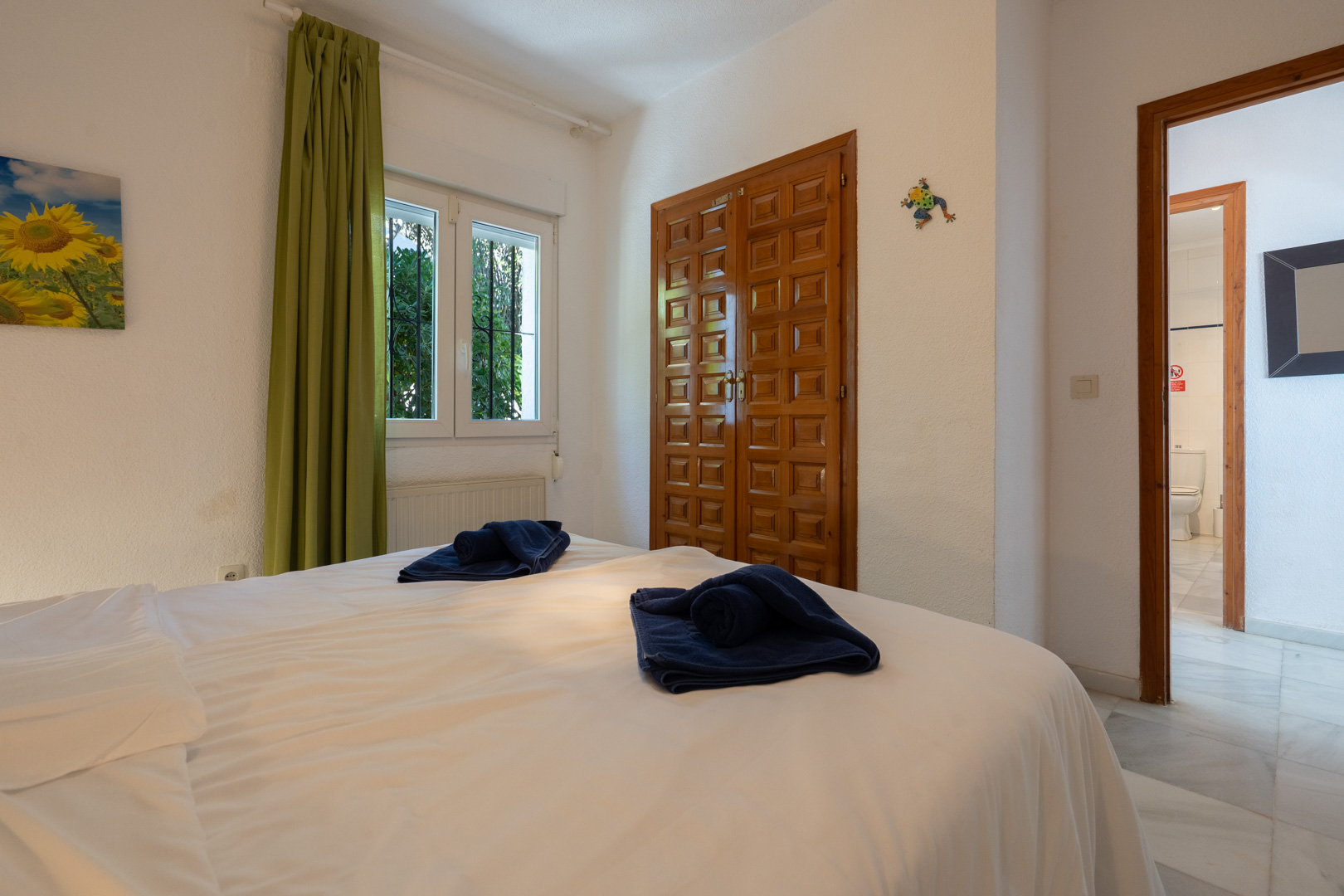 Dormitorio luminoso con armario empotrado de madera tallada, ventana con cortinas verdes y acceso visual a un baño con suelos de mármol.
