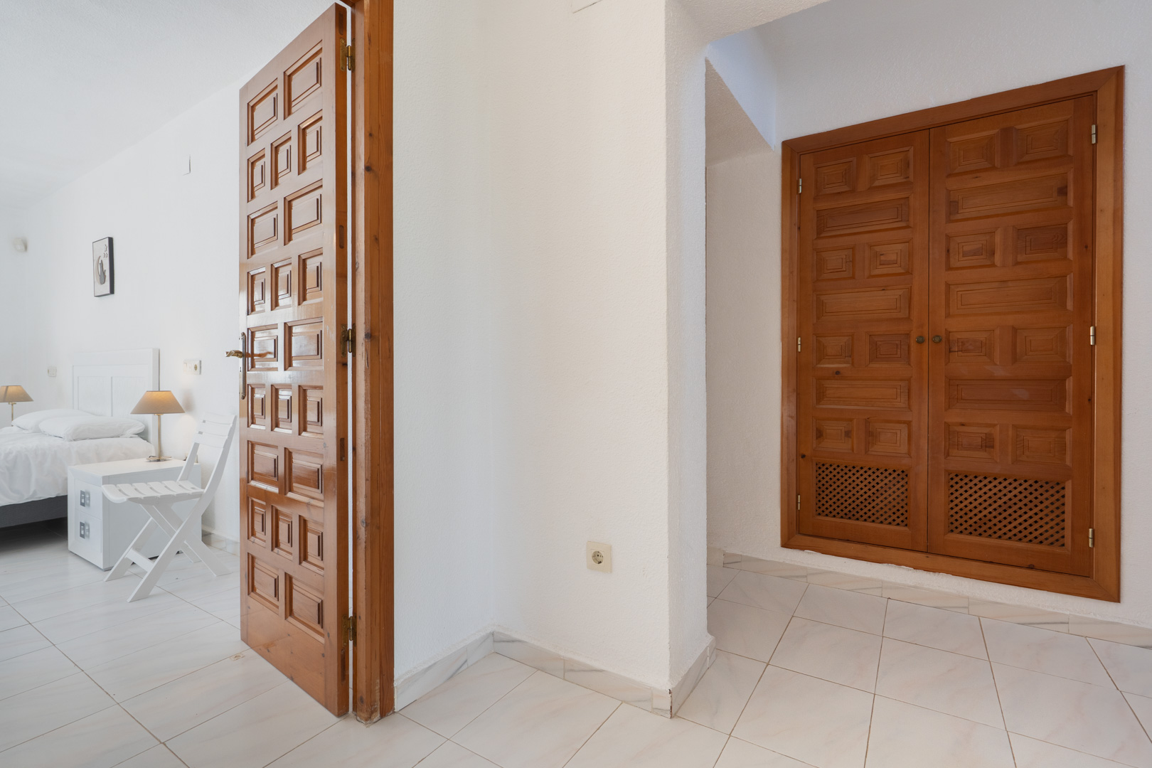 Pasillo con puertas de madera tallada de estilo tradicional y armarios empotrados, que da acceso a un dormitorio luminoso con suelos cerámicos blancos.
