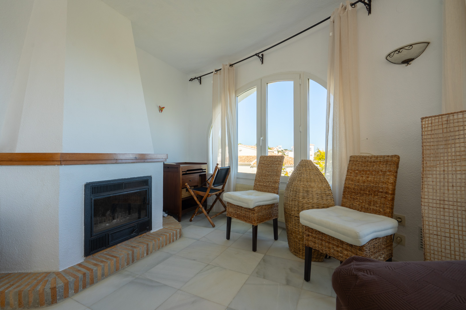 Salón con chimenea de obra con base de ladrillo, suelos de mármol y ventanal arqueado que aporta luz natural a la zona de estar con mobiliario de mimbre.