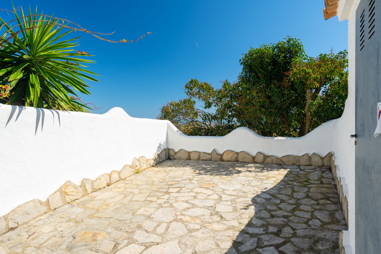 Terraza exterior con suelo de piedra natural irregular y muros blancos de remate ondulado, integrada en un entorno con vegetación mediterránea.