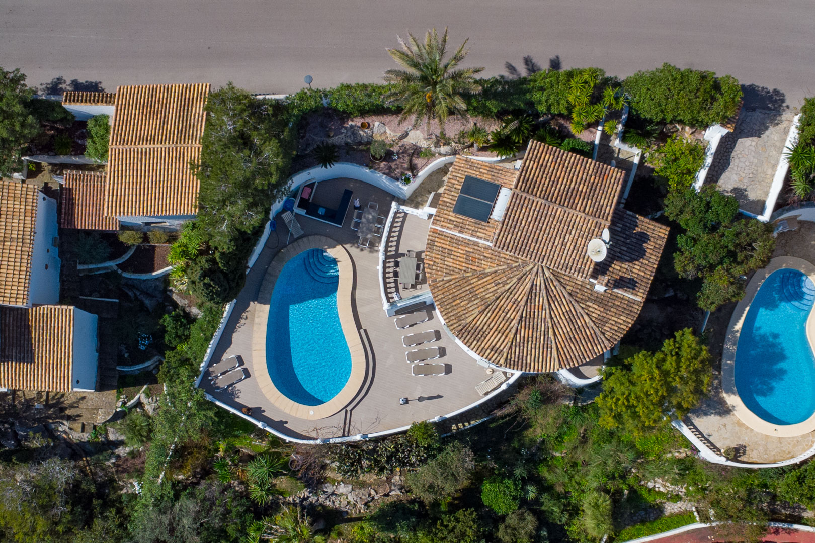 Vista aérea de la propiedad que destaca la piscina de forma curva, el amplio solárium con comedor exterior y el tejado tradicional con placas solares.