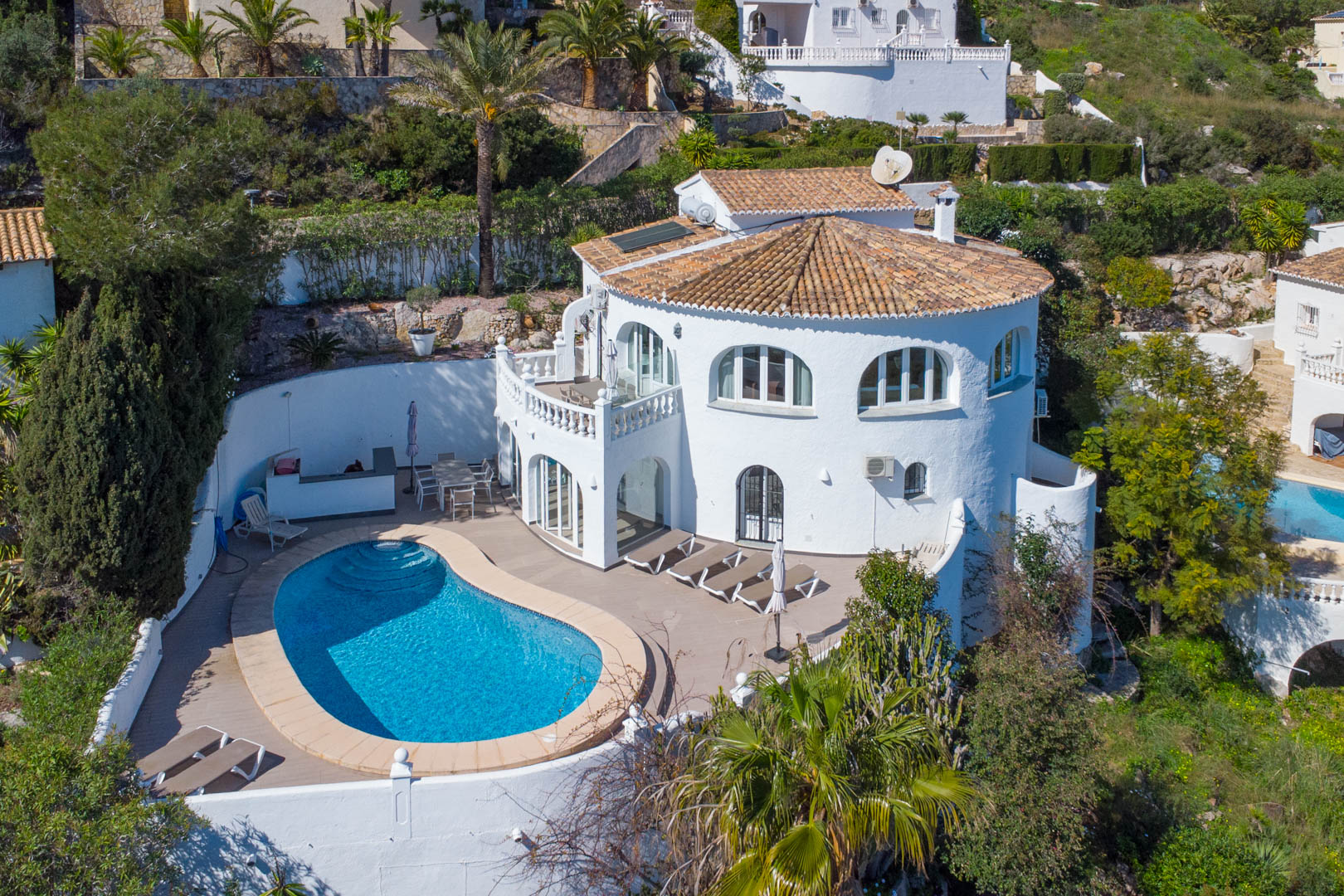 Vista aérea de la villa con piscina en forma de riñón, amplia terraza solárium, fachada blanca tradicional y paneles solares sobre el tejado de teja.