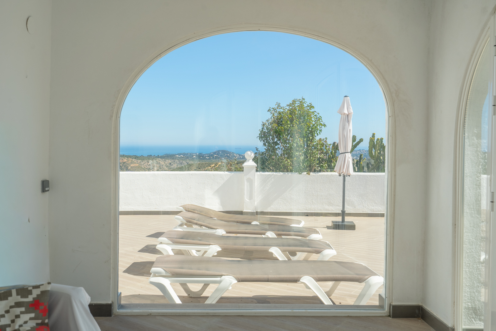 Vista desde la naya acristalada a través de un gran arco, mostrando la terraza solárium con tumbonas y vistas panorámicas al mar Mediterráneo.