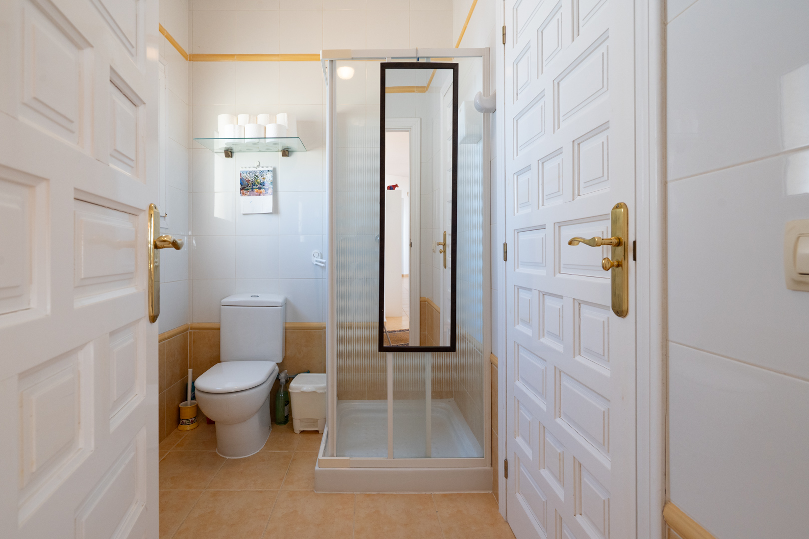 Baño funcional con azulejos blancos y ocre, equipado con inodoro y cabina de ducha con espejo, accesible a través de puertas blancas de paneles.