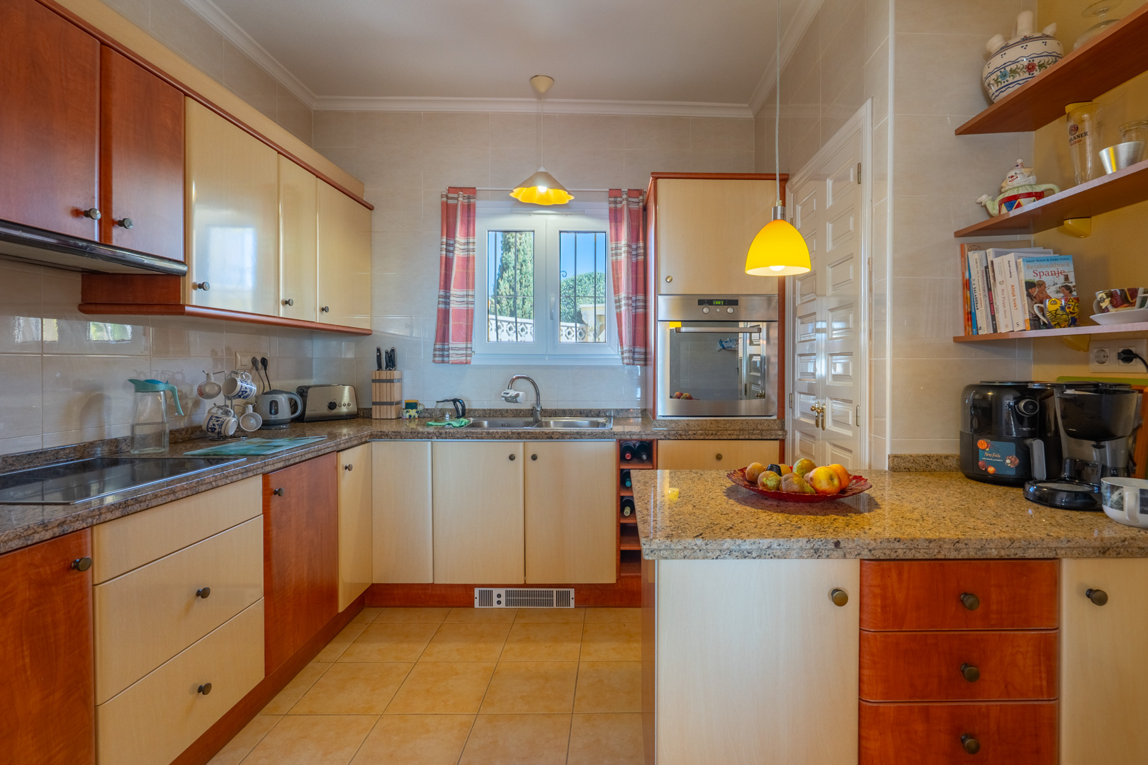 Cocina equipada con encimeras de granito, armarios bitono en madera y crema, y electrodomésticos integrados. Incluye estanterías abiertas y una ventana que aporta luz natural.