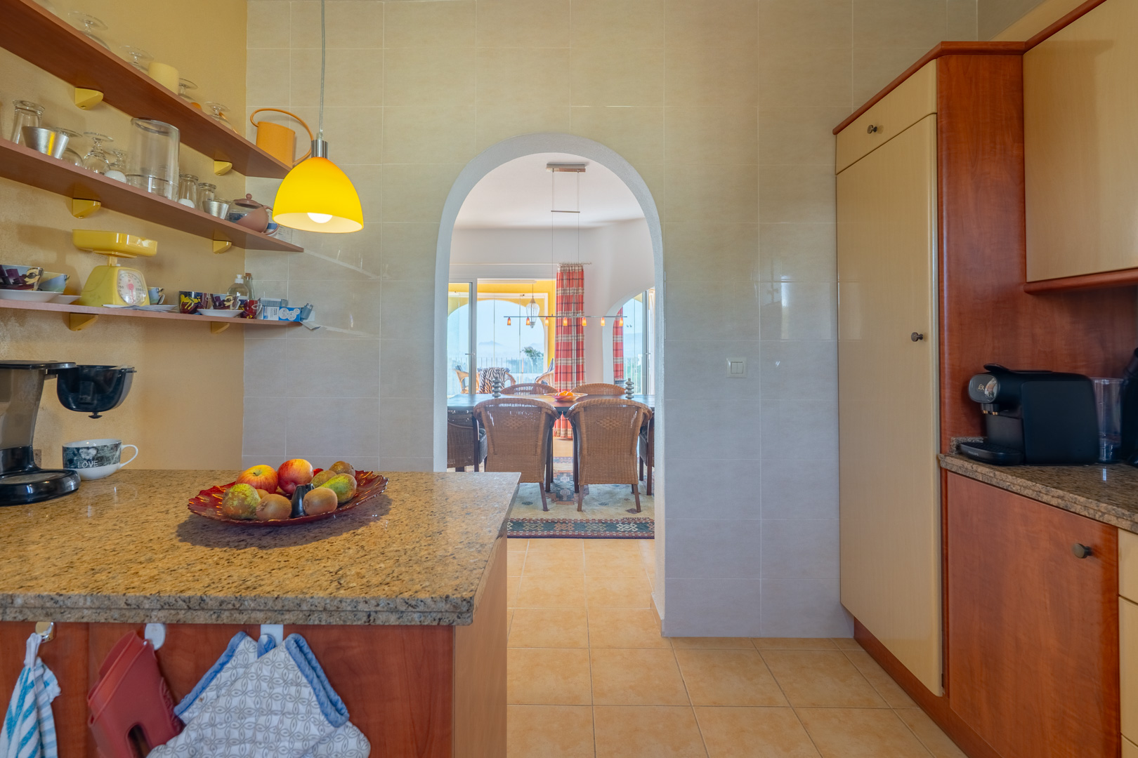Cocina funcional con encimeras de granito y estanterías abiertas, comunicada mediante un arco con el comedor y la terraza exterior.