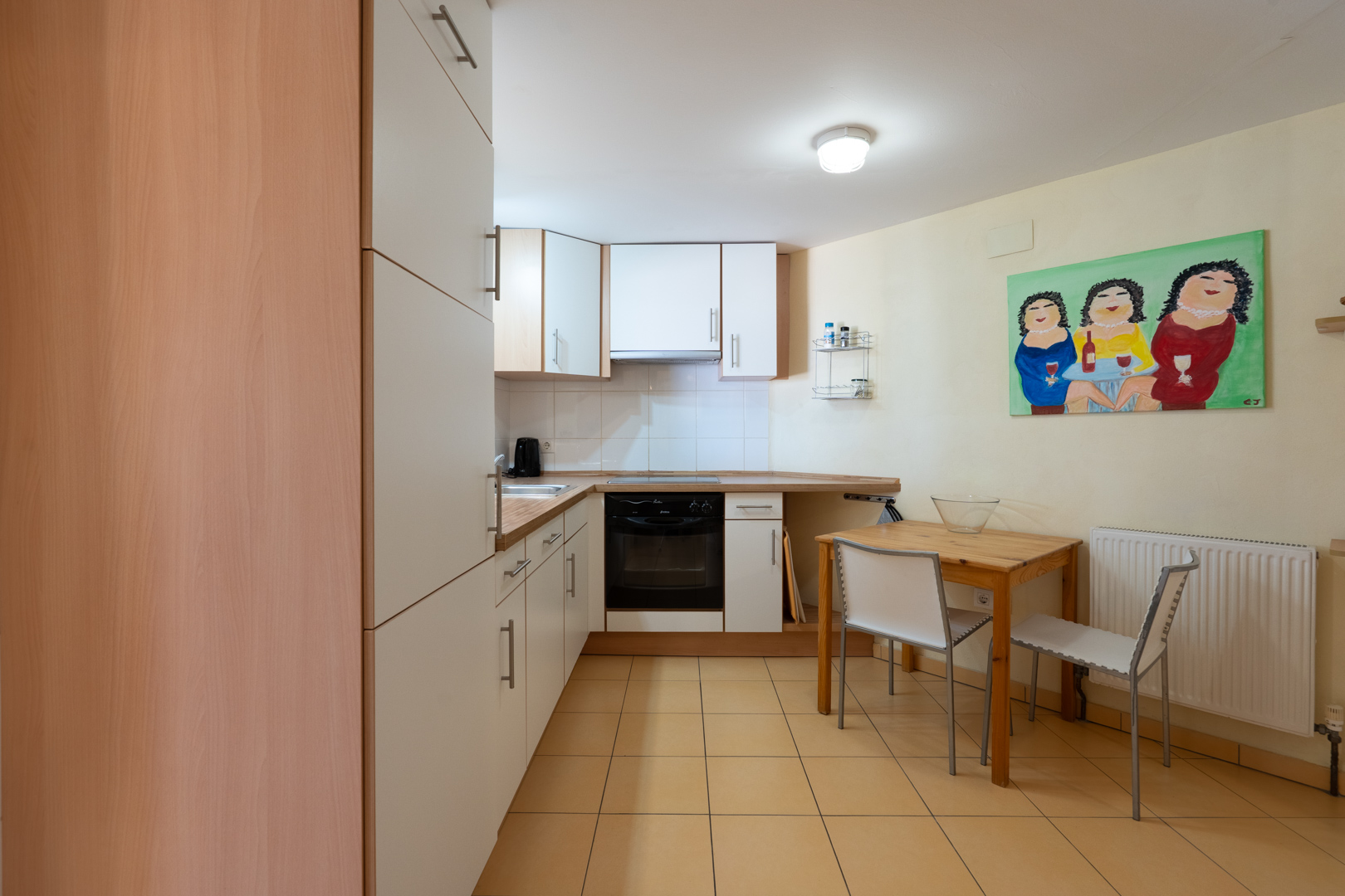 Cocina funcional del apartamento de invitados con armarios blancos, encimeras de madera, zona de comedor compacta y suelos cerámicos en tonos cálidos.