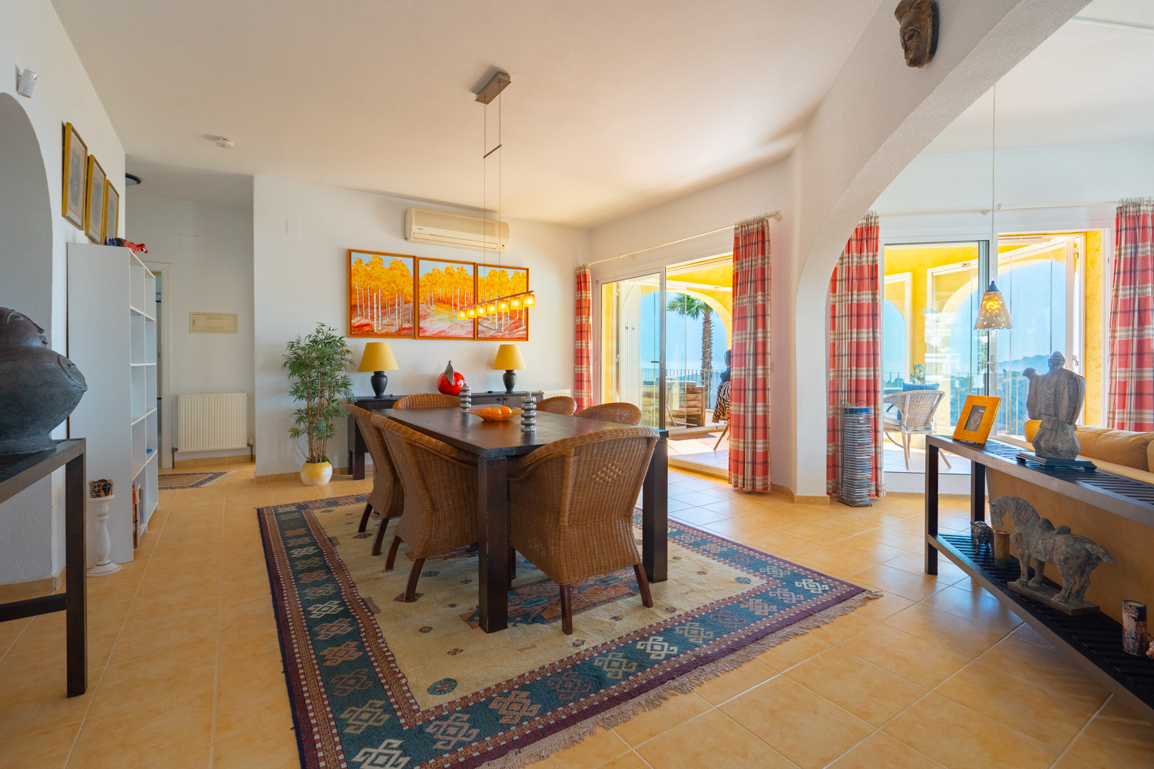 Comedor con mesa de madera oscura y sillas de mimbre sobre suelo cerámico, con acceso directo a la naya arqueada y vistas al mar a través de ventanales.
