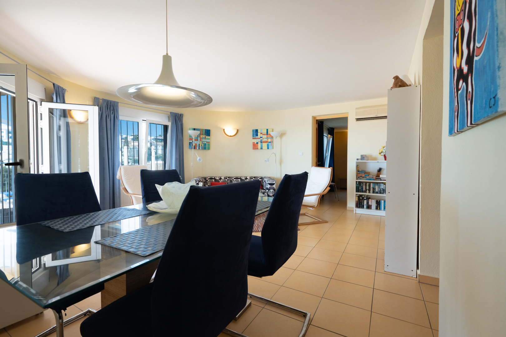 Comedor luminoso con mesa de cristal y sillas de terciopelo azul, integrado en un salón de planta abierta con suelos cerámicos y acceso directo al exterior.
