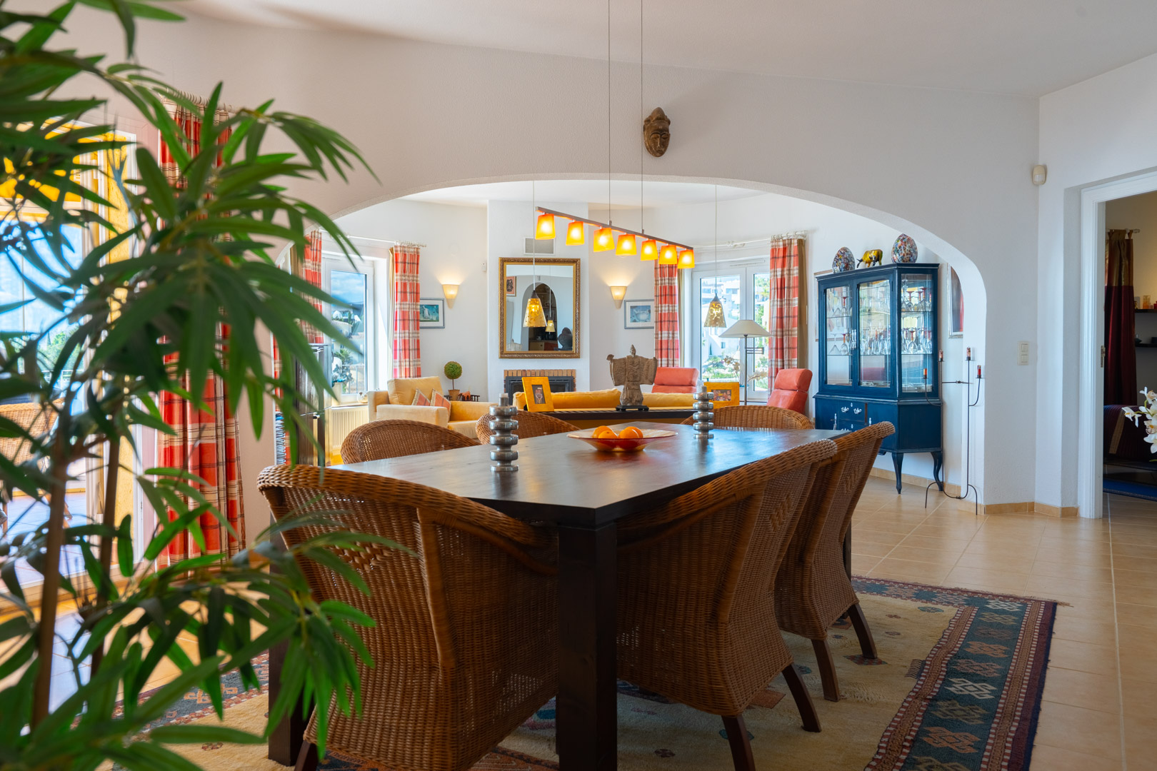 Comedor luminoso con mesa de madera oscura y sillas de mimbre, conectado al salón mediante un arco. Incluye suelos de gres y abundante luz natural.