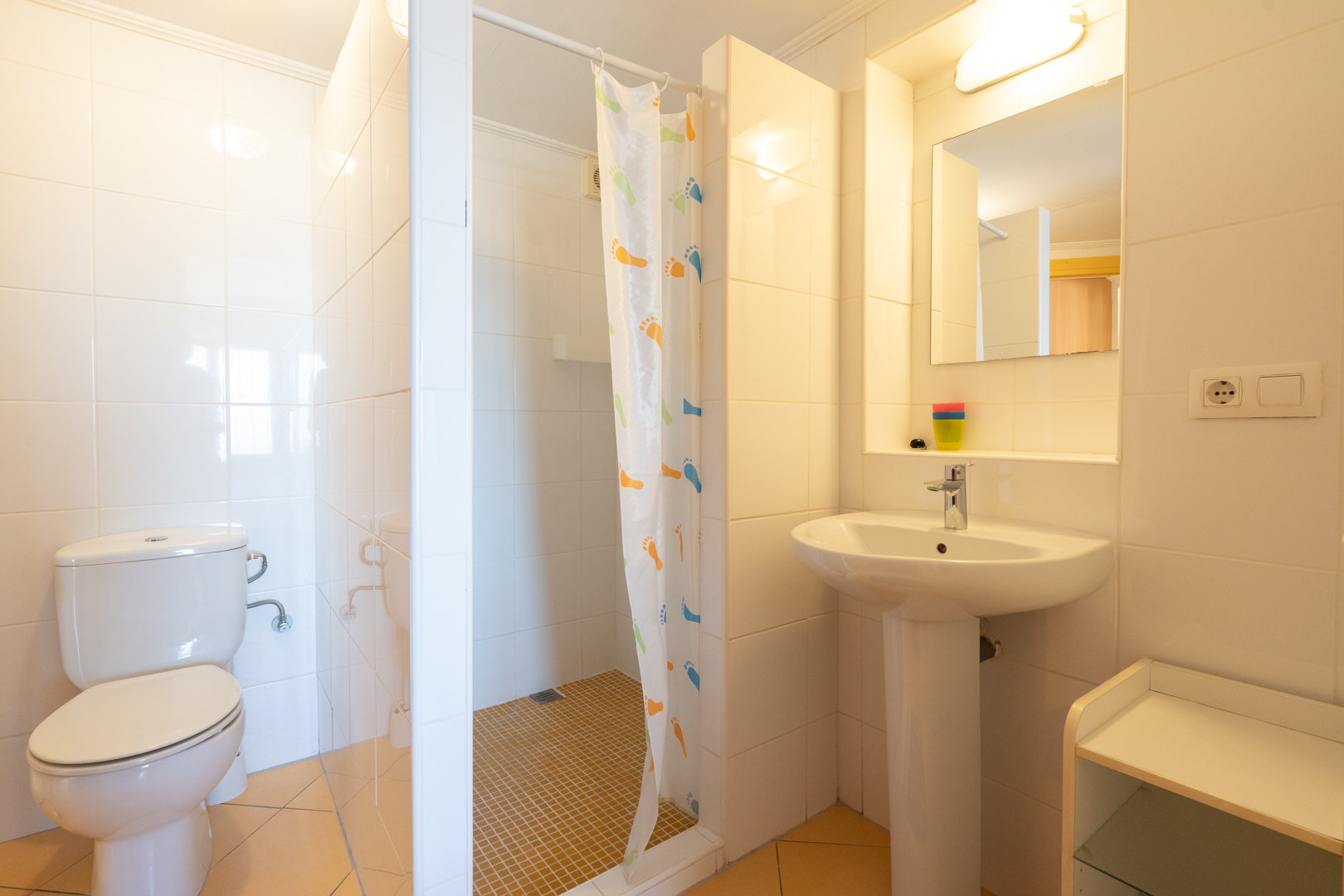 Cuarto de baño funcional con azulejos blancos, ducha de obra con cortina decorativa, lavabo de pedestal y sanitario clásico en tonos claros.