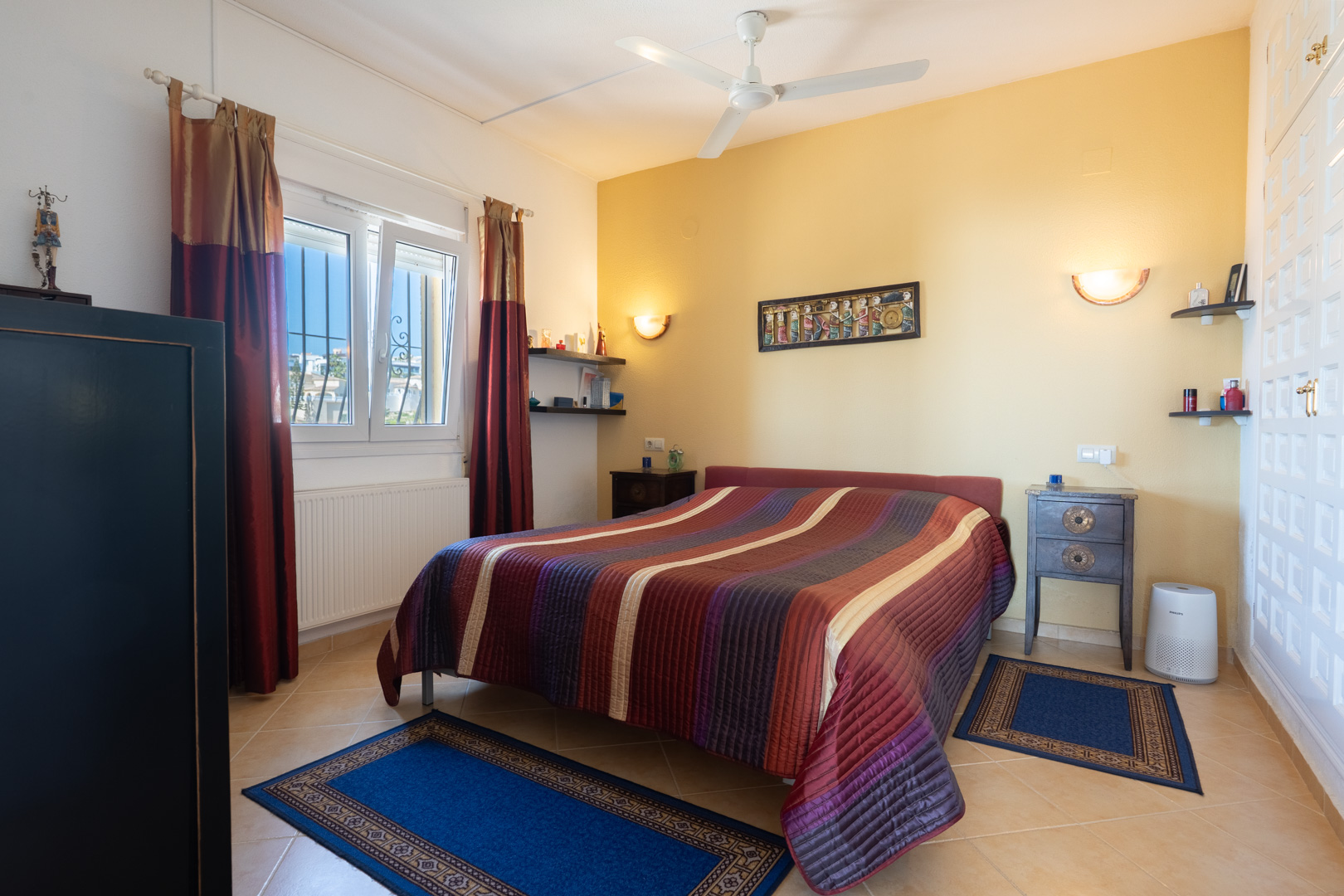 Dormitorio con cama doble, pared de acento amarilla, ventilador de techo y armarios empotrados blancos. Incluye calefacción por radiador y suelos cerámicos.