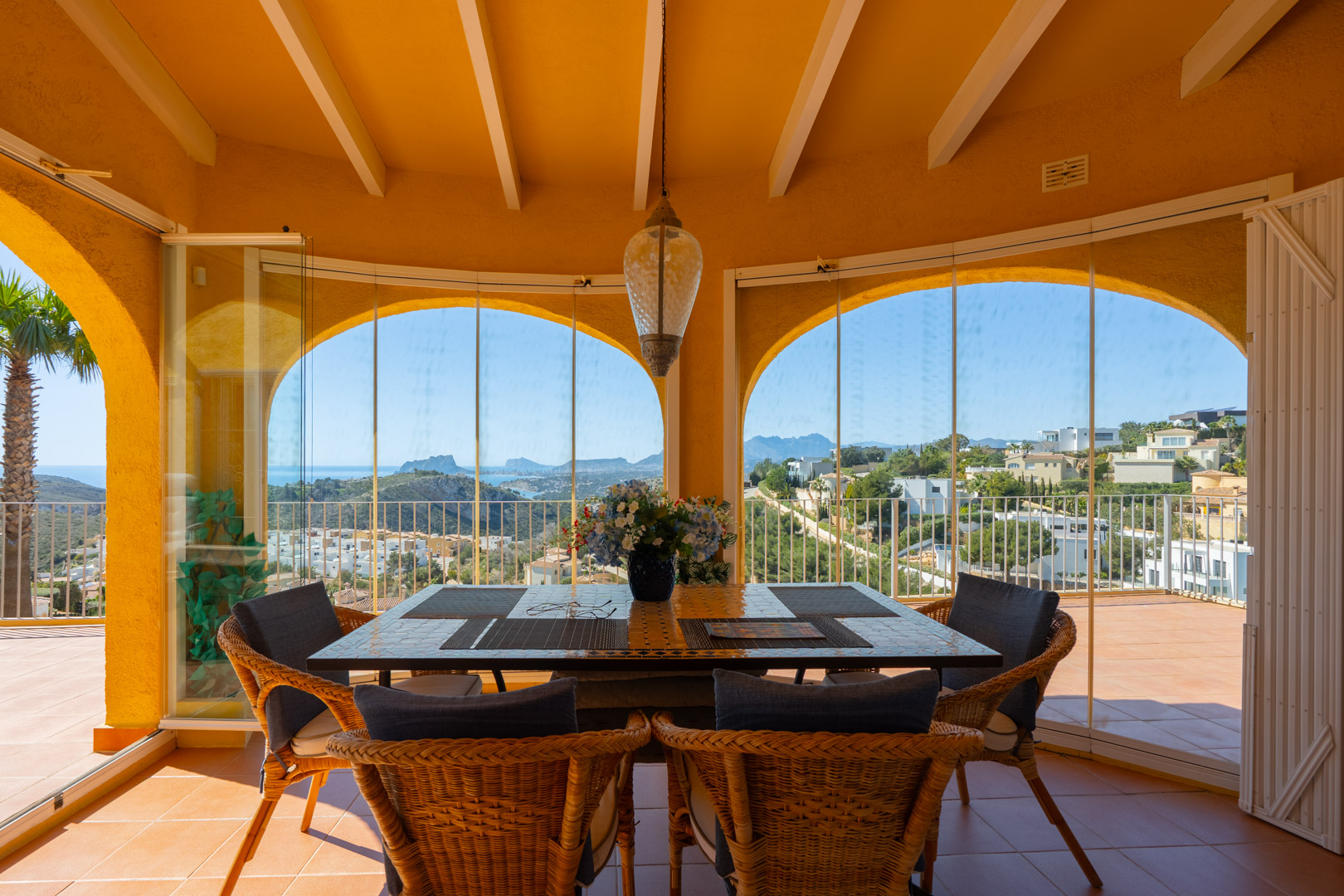 Naya acristalada con arcos tradicionales y vigas de madera, que ofrece vistas panorámicas al mar y las montañas desde el comedor exterior con suelo de terracota.