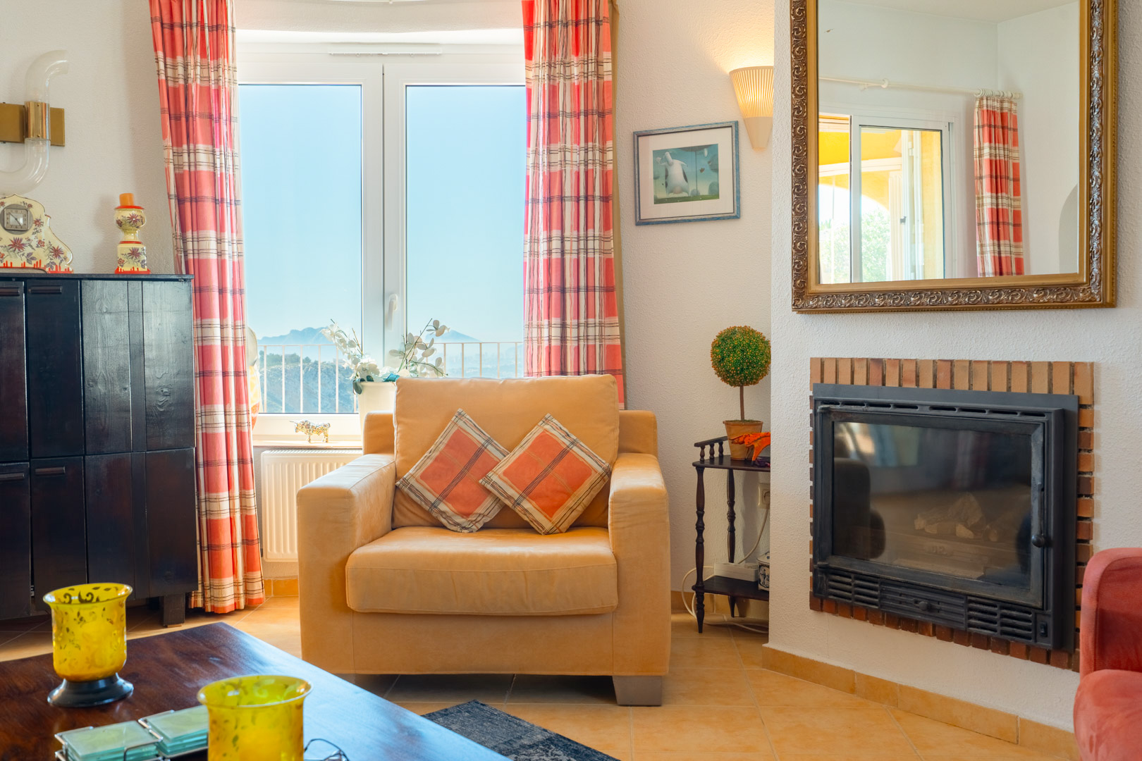 Salón con chimenea encastrada de ladrillo, sillón tapizado y ventanal con vistas a las montañas. Incluye un gran espejo con marco dorado y suelos cerámicos.