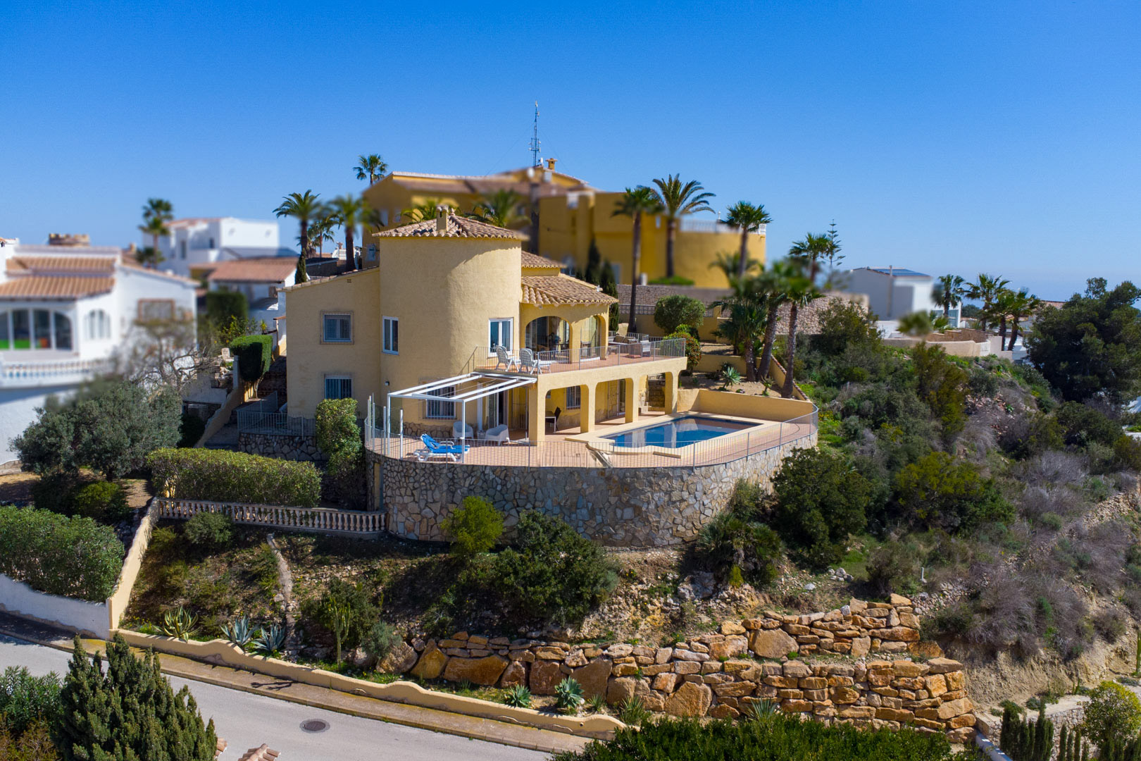 Vista exterior de la villa con fachada ocre, torre circular y piscina rectangular integrada en una amplia terraza con muros de piedra natural.