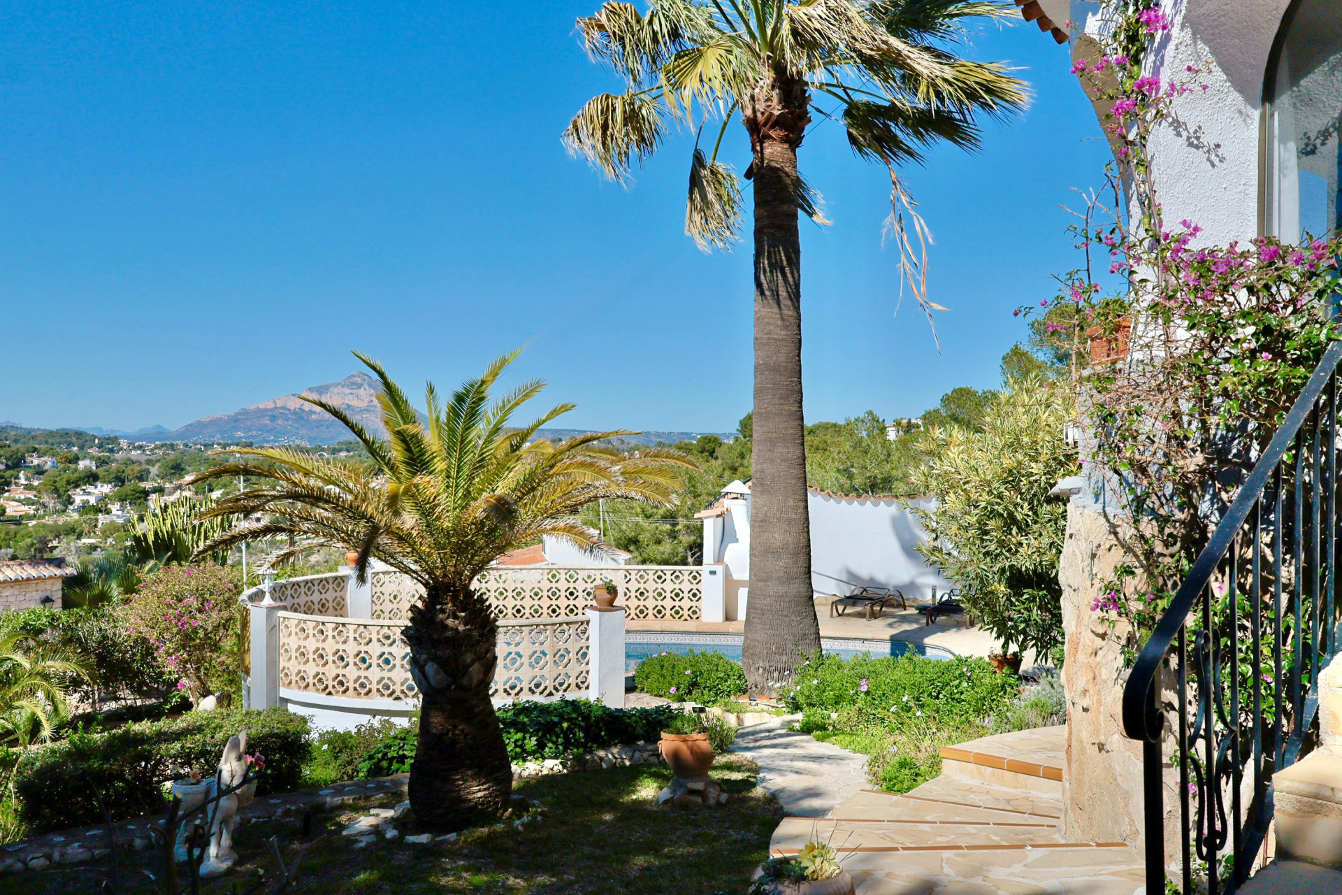 Vista del jardín con piscina y palmeras, destacando la vegetación mediterránea y el paisaje montañoso al fondo.