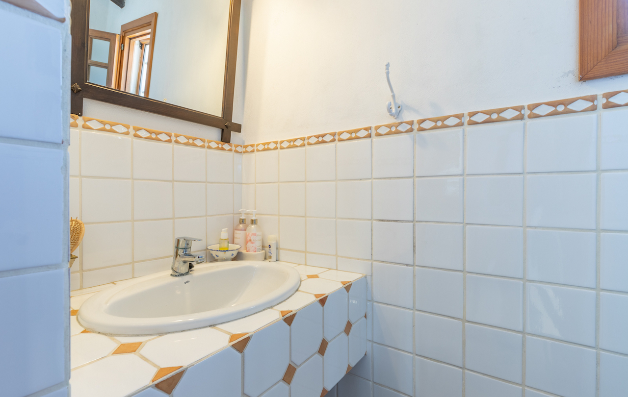 Baño de estilo tradicional con azulejos blancos y cenefa decorativa. Lavabo encastrado sobre encimera alicatada con detalles geométricos y espejo con marco de madera.