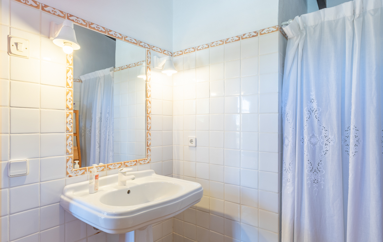 Baño de estilo tradicional con lavabo de pedestal blanco, espejo con cenefa cerámica decorativa y apliques de pared clásicos junto a una cortina bordada.