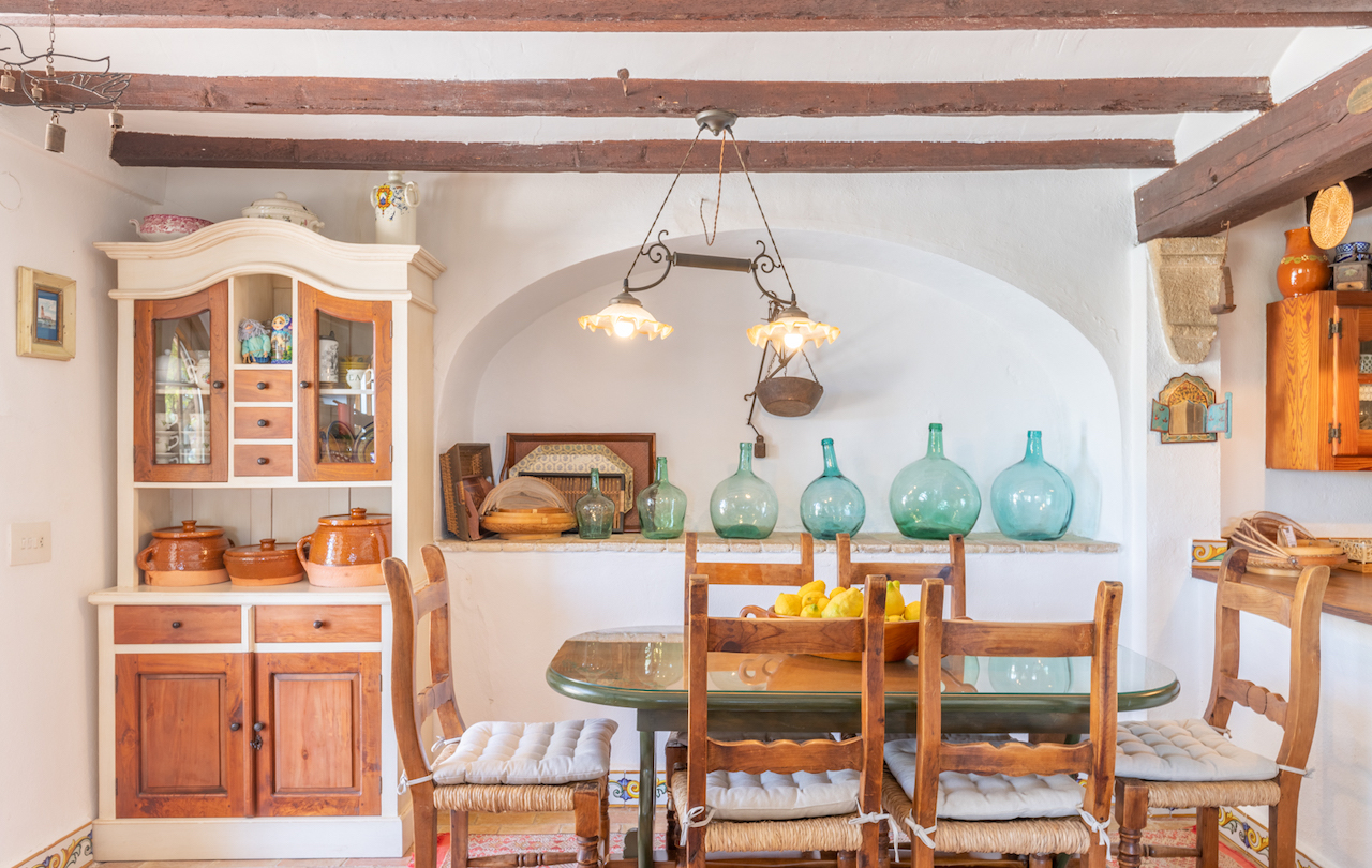 Comedor de estilo rústico valenciano con vigas de madera vista, alacena tradicional y hornacina decorativa con damajuanas de cristal sobre el conjunto de mesa y sillas.