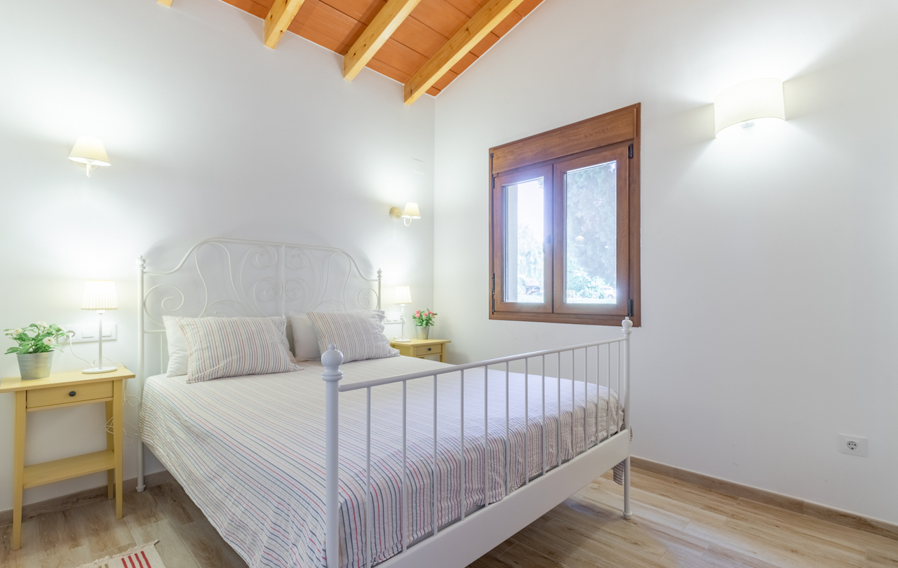 Dormitorio rústico con techo de vigas de madera y bovedillas, cama de forja blanca y ventana de madera con luz natural.