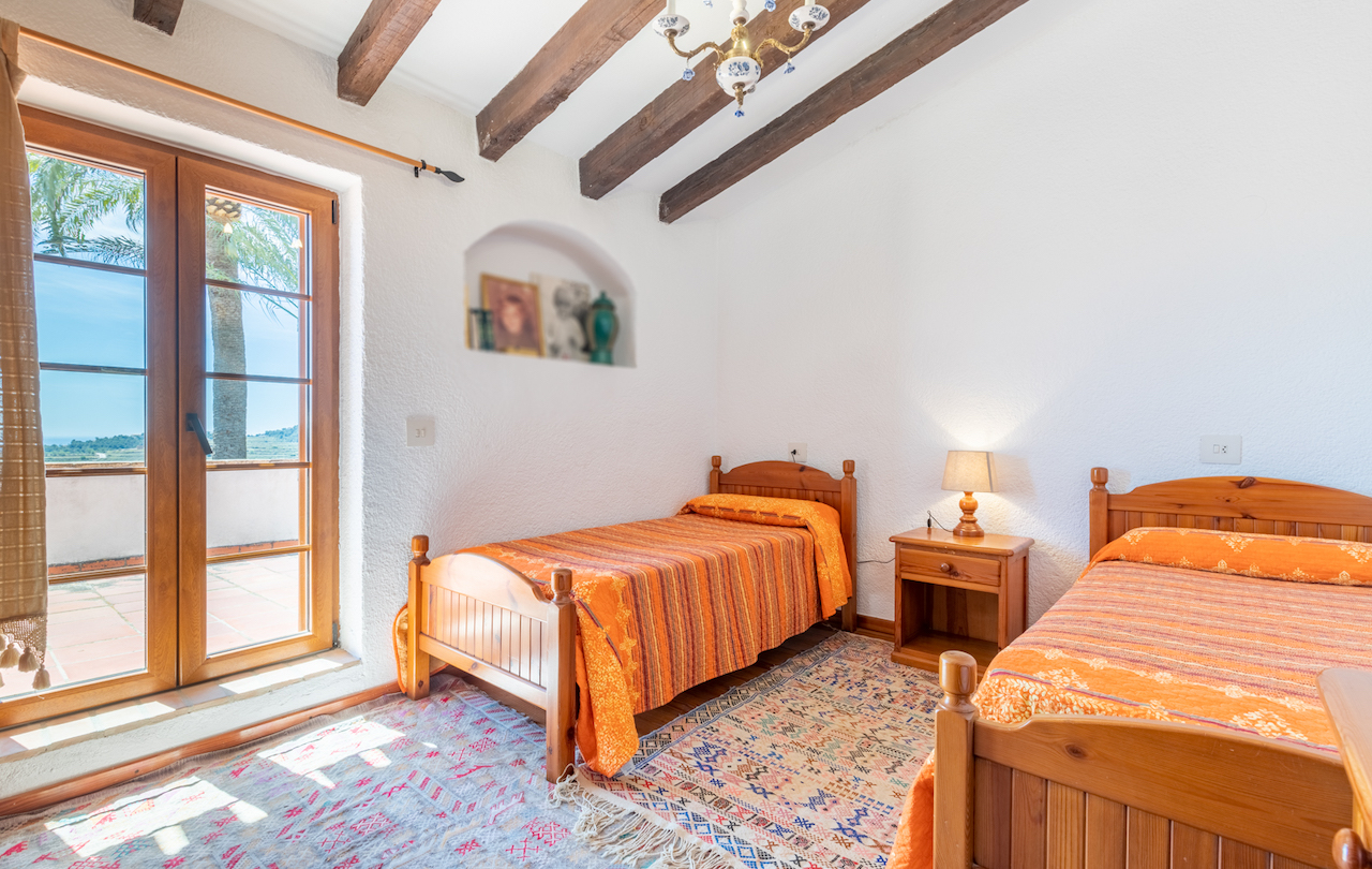 Dormitorio rústico con vigas de madera vista, dos camas individuales y acceso directo a la terraza. Destaca el nicho decorativo en la pared y la abundante luz natural.