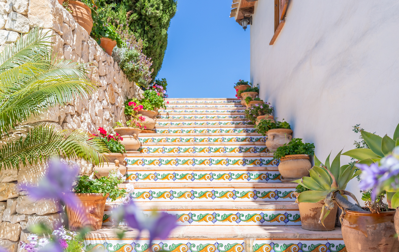 Escalera exterior de estilo tradicional con contrahuellas decoradas con azulejos pintados a mano, flanqueada por macetas de terracota y un muro de piedra natural.