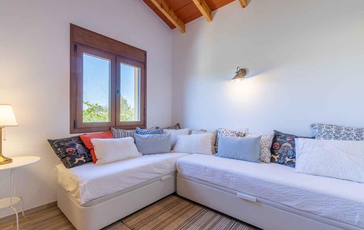 Habitación con sofá cama en L, techos de vigas de madera vista y carpintería de madera con vistas al exterior. Decoración con textiles variados y luz cálida.