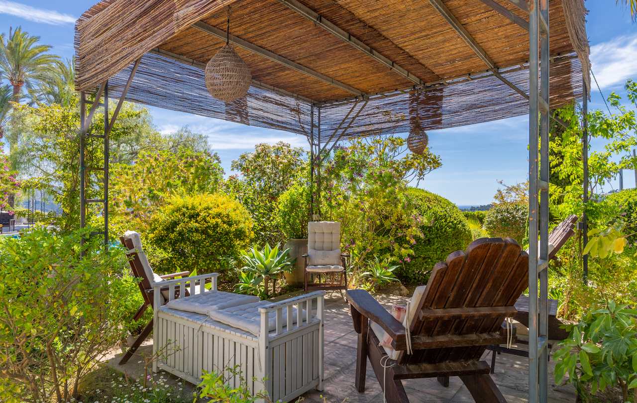Pérgola de estructura metálica y techado de cañizo con zona de estar, rodeada de vegetación mediterránea y vistas despejadas hacia la costa.