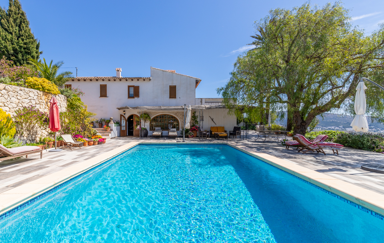 Piscina rectangular rodeada de una amplia terraza de gres antideslizante, con vistas a la fachada tradicional blanca y un jardín maduro con muros de piedra natural.
