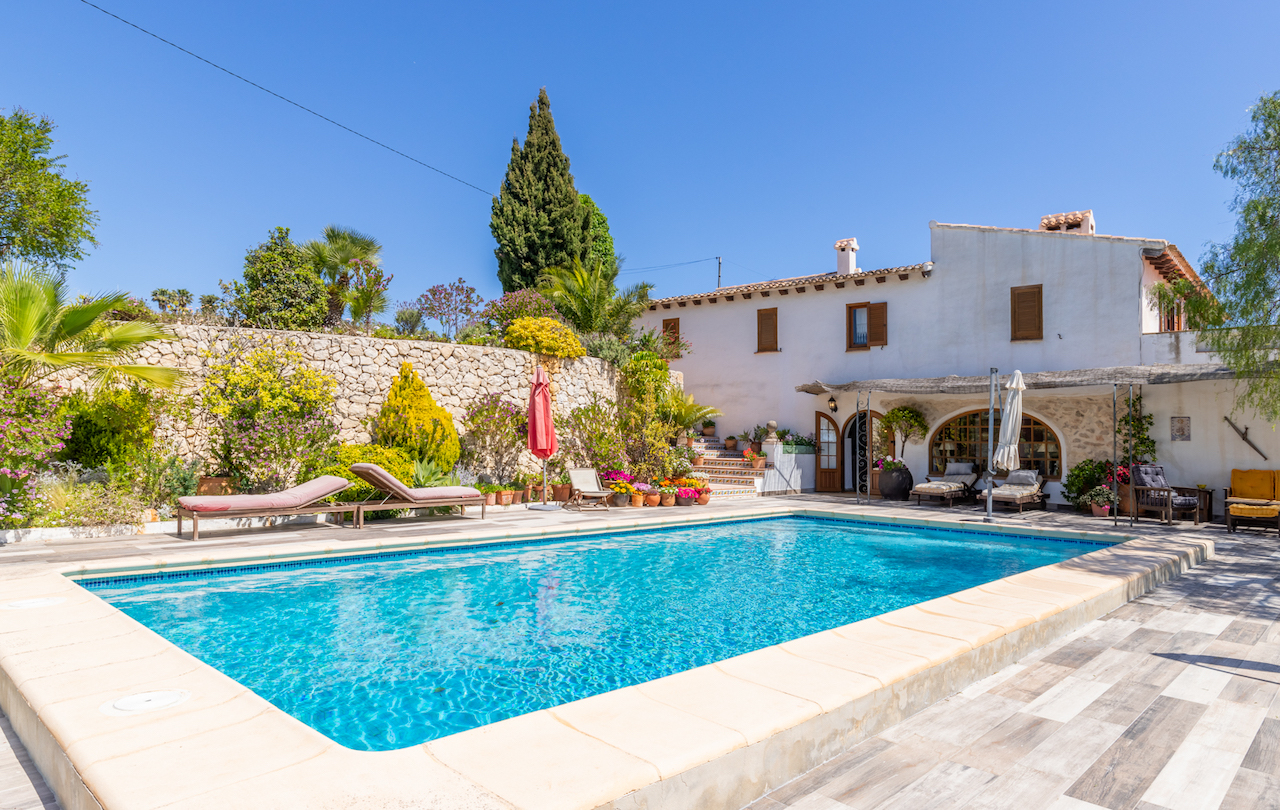 Piscina rectangular rodeada de una amplia terraza de pavimento antideslizante, muros de piedra natural y vegetación mediterránea junto a la fachada de estilo tradicional.