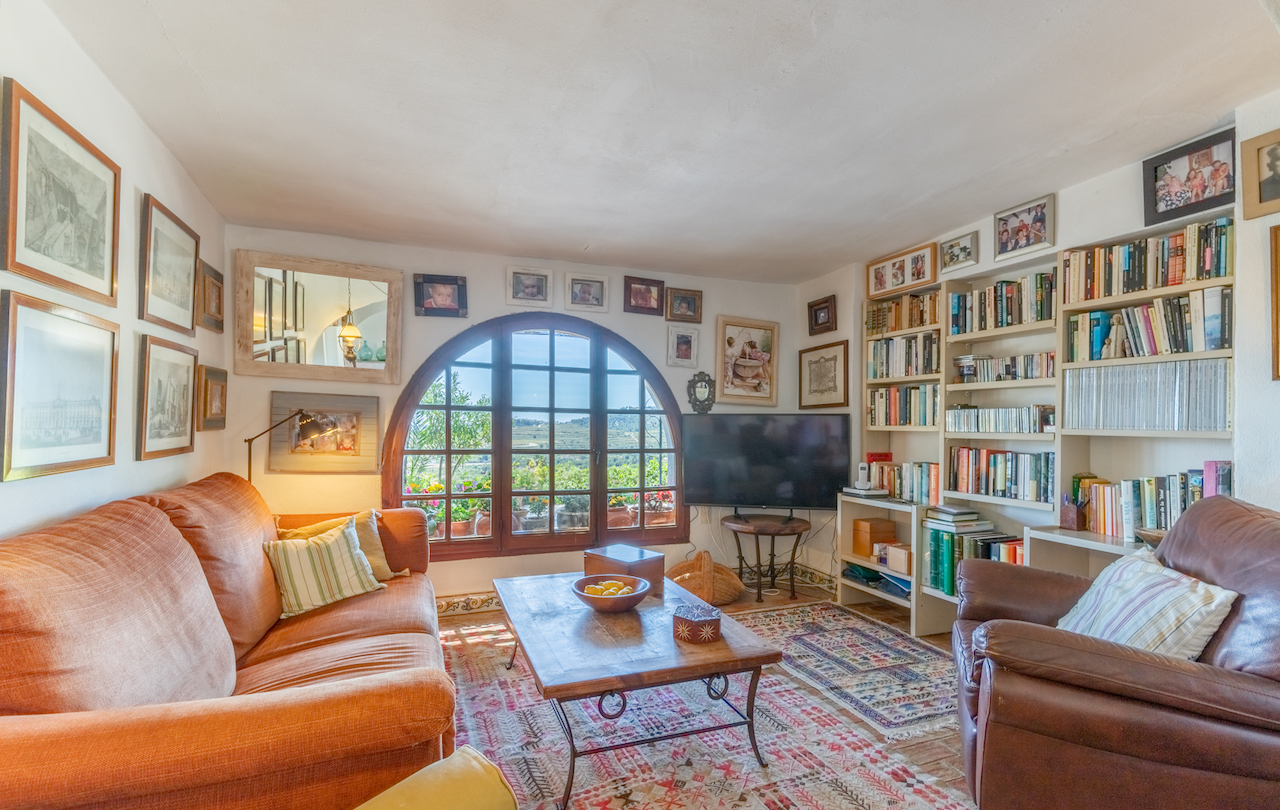 Salón biblioteca con ventanal de medio punto de madera, estanterías integradas, suelos de barro con alfombras tradicionales y vistas despejadas al entorno rural.