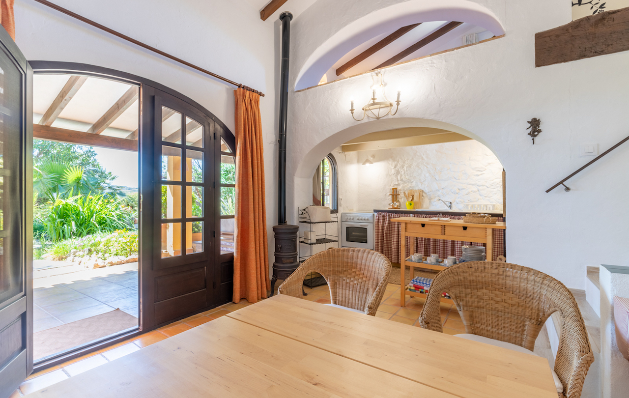 Salón comedor rústico con vigas de madera, estufa de hierro y suelos de terracota. La cocina integrada se enmarca en un arco tradicional con acceso directo al jardín exterior.