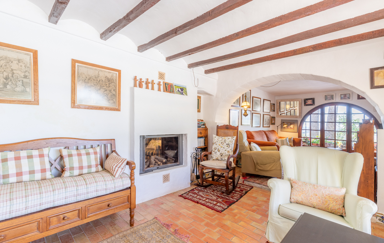 Salón tradicional con vigas de madera vistas, suelos de terracota y una chimenea encastrada. Un arco conecta con una zona de estar luminosa junto a un ventanal de medio punto.