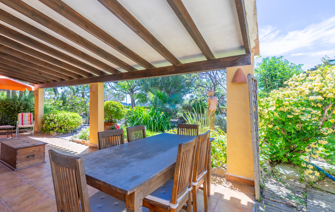 Terraza cubierta con vigas de madera oscura y suelos de gres, equipada con comedor exterior frente a un frondoso jardín mediterráneo con vegetación madura.