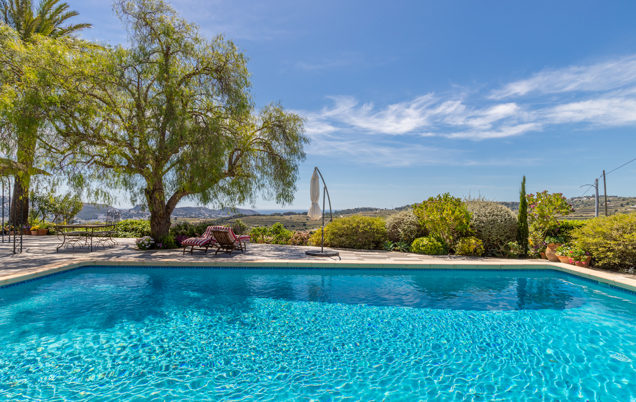 Terraza de más de 200m2 con piscina, zona de descanso bajo árboles maduros y vistas abiertas a los campos de viñedos y al mar de Moraira.