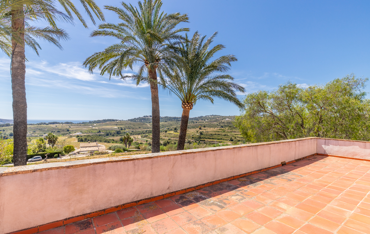 Terraza elevada con suelo de baldosas de barro, ofreciendo vistas panorámicas a los campos de viñedos aterrazados y al mar Mediterráneo bajo un cielo despejado.
