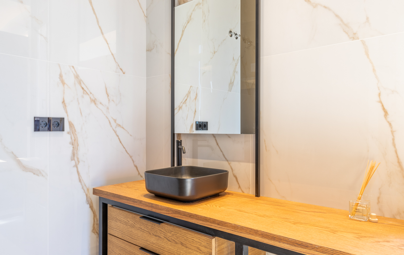Detalle de baño con lavabo negro mate sobre encimera de madera clara y pared de mármol crema veteado.