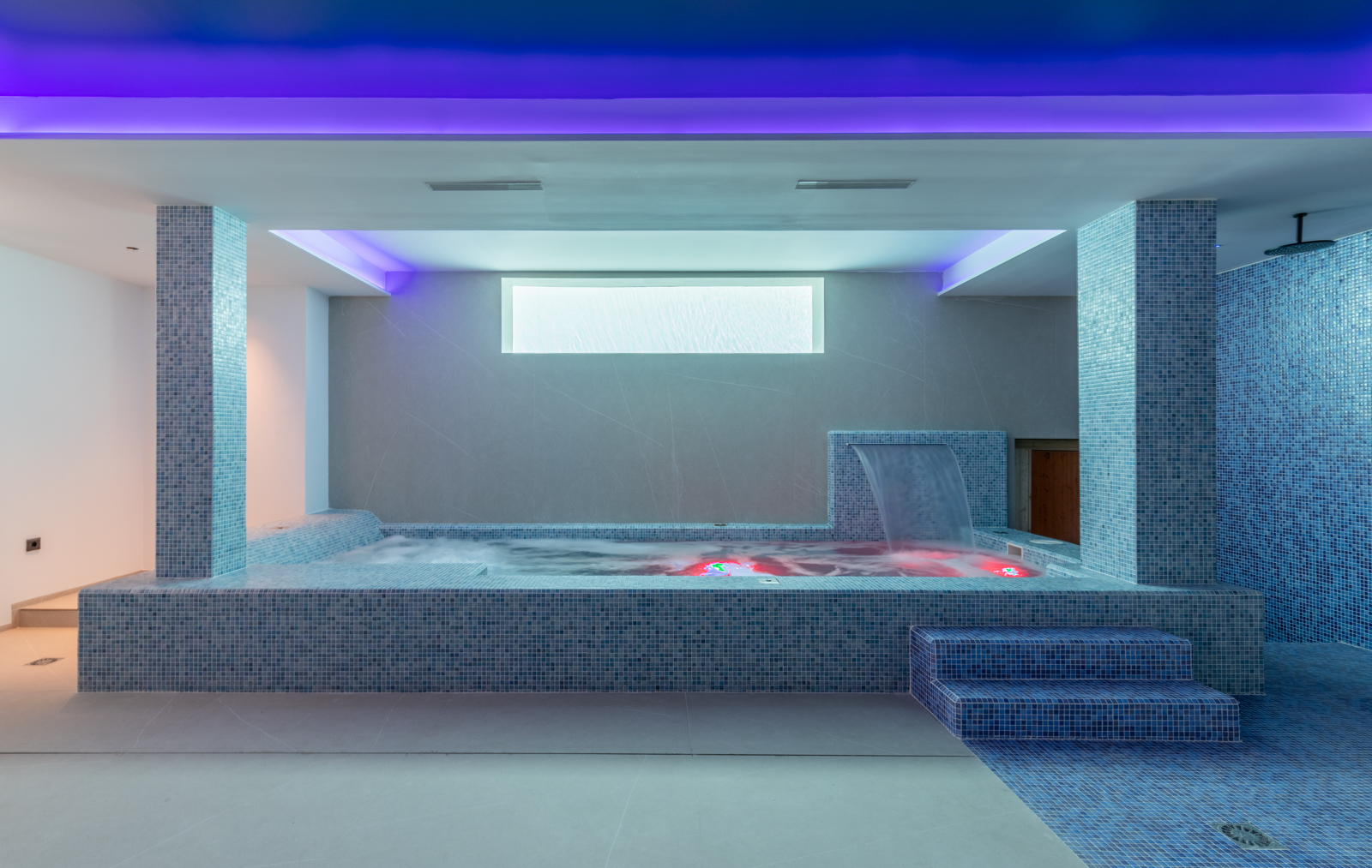 Detalle del spa interior con jacuzzi, cascada y ducha con mosaicos azules e iluminación ambiental.