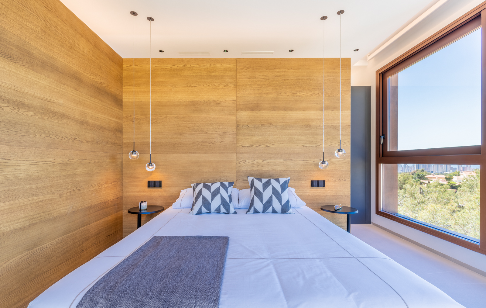 Dormitorio con pared de madera clara, lámparas colgantes esféricas y vistas al exterior.