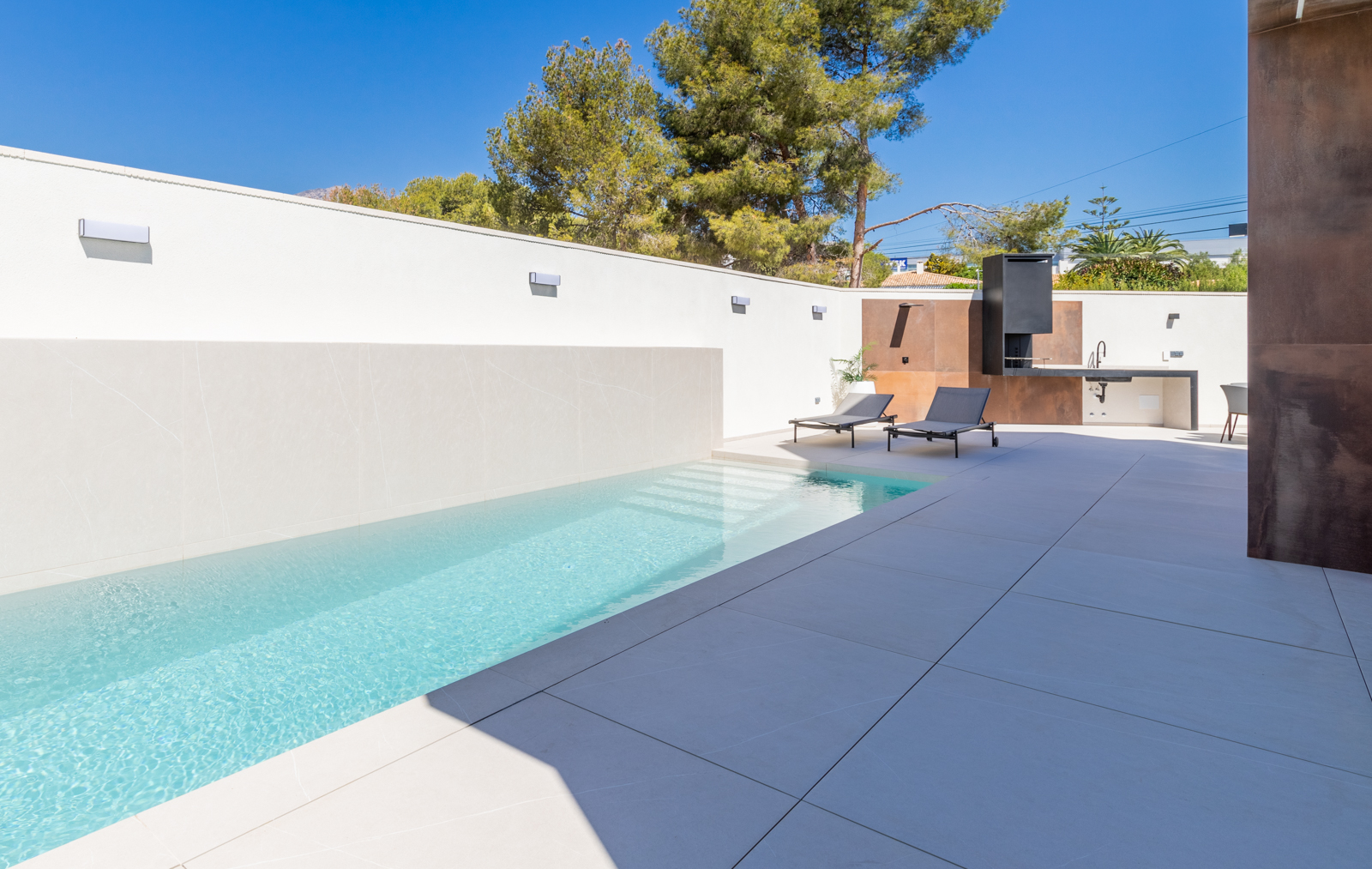 Piscina moderna con pared de azulejos blancos y zona de barbacoa con encimera de hormigón.