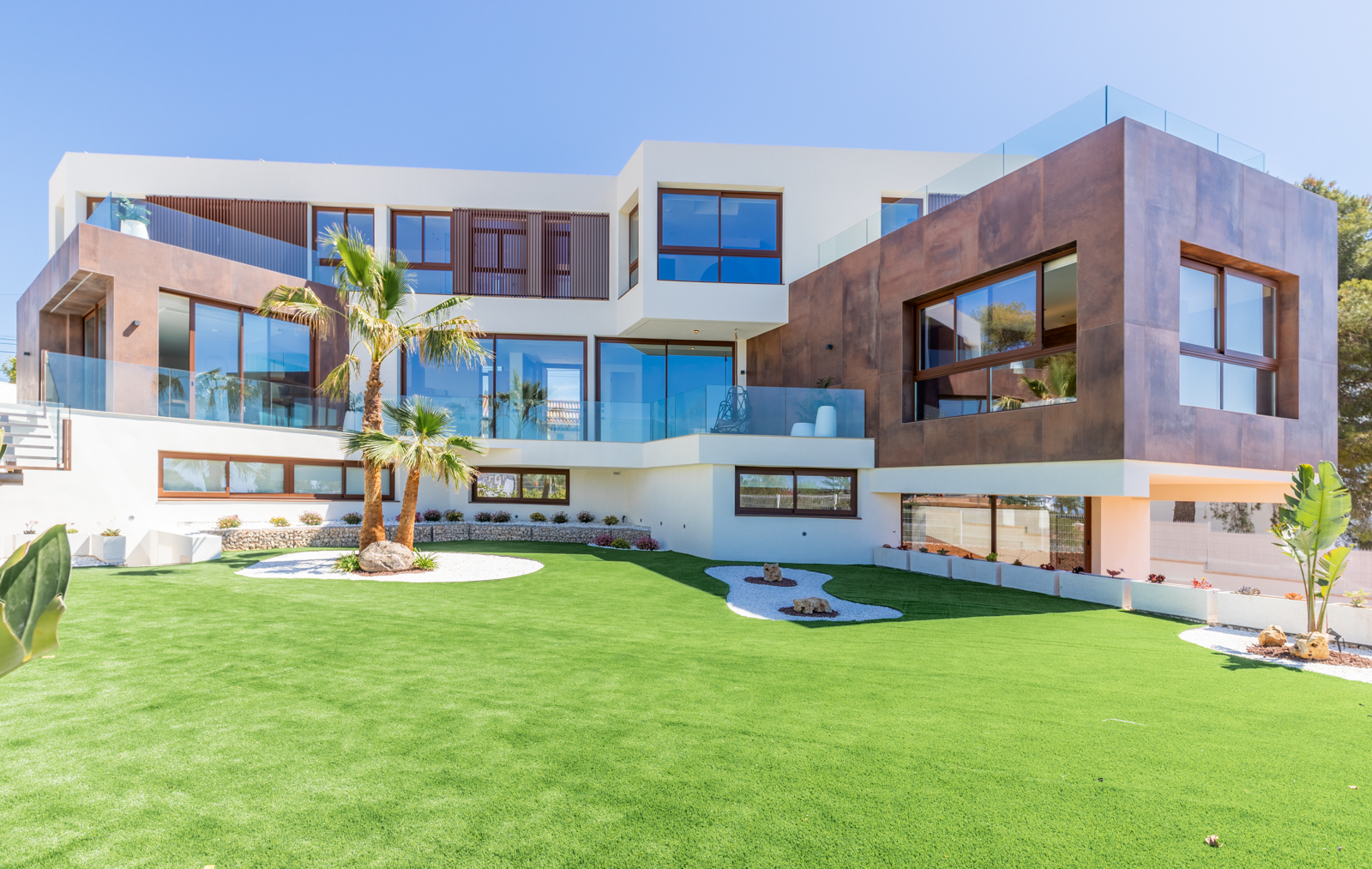 Vista exterior de la villa con jardín de césped, palmeras y fachada moderna con grandes ventanales.