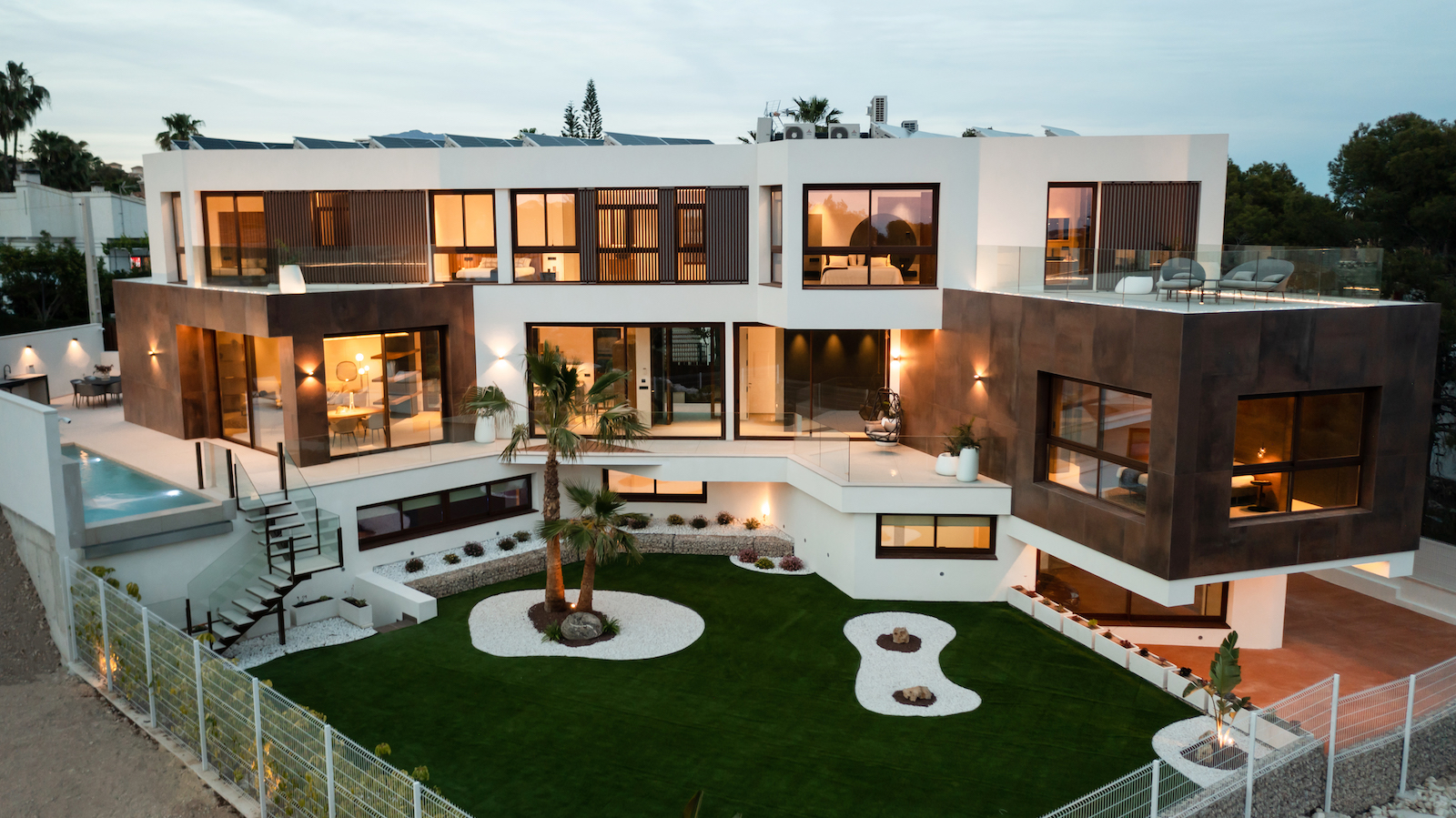 Vista exterior de la villa moderna con jardín mediterráneo, piscina y terrazas iluminadas.