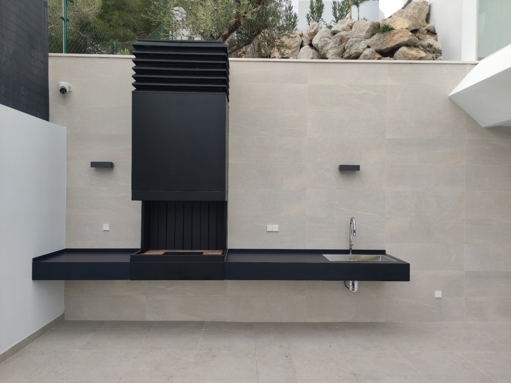 Cocina exterior de diseño minimalista en negro mate, equipada con barbacoa integrada, fregadero de acero inoxidable y encimera sobre pared de piedra natural.