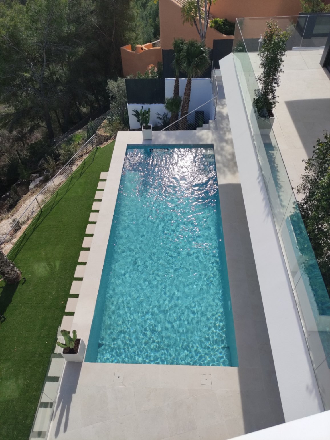Piscina rectangular de diseño minimalista con revestimiento azul, rodeada por una terraza de piedra clara, césped artificial y barandillas de cristal de seguridad.