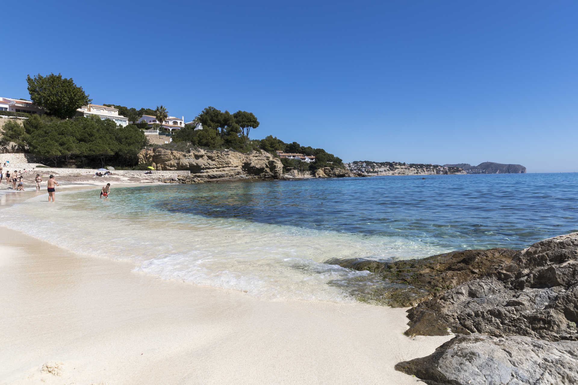 Playa de arena blanca y aguas cristalinas en La Fustera, rodeada de formaciones rocosas y vegetación mediterránea bajo un cielo despejado.