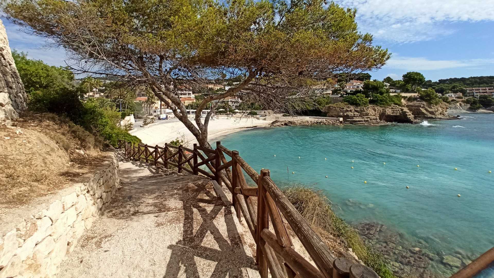Sendero costero con barandilla de madera y muros de piedra natural que conduce directamente a la playa de arena de La Fustera y sus aguas cristalinas.