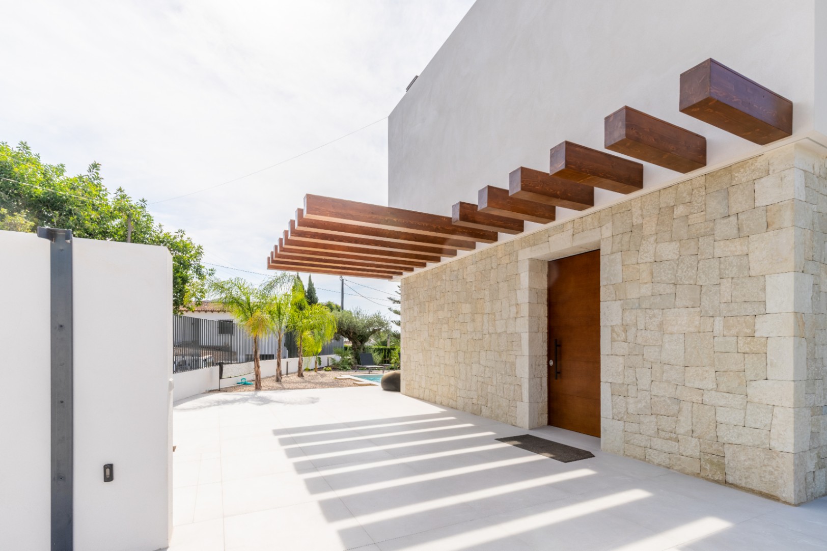 Entrada principal con pared de piedra y pérgola de madera que proyecta sombras sobre el suelo.